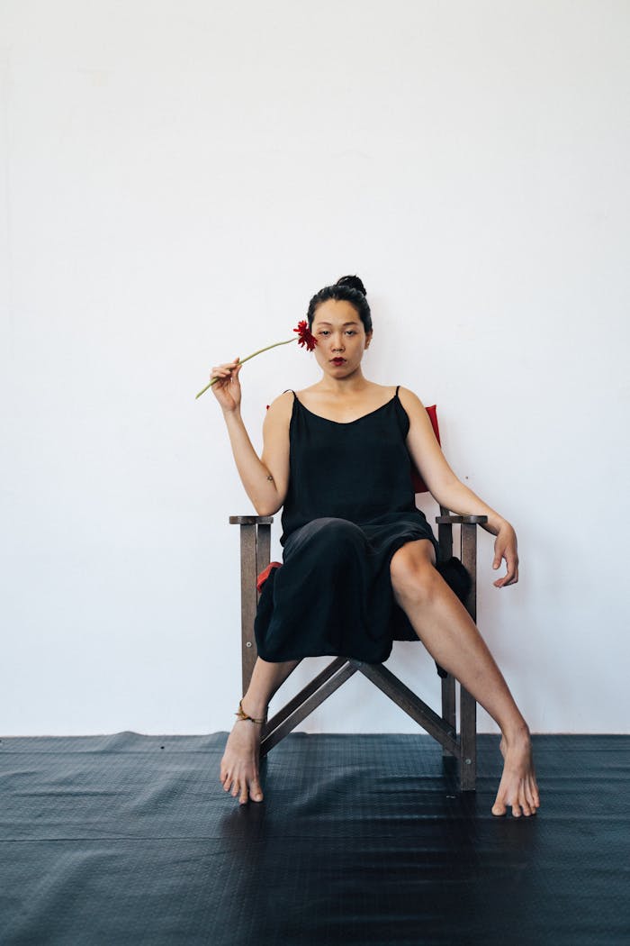 Asian woman in black dress with red flower, sitting barefoot on a chair.